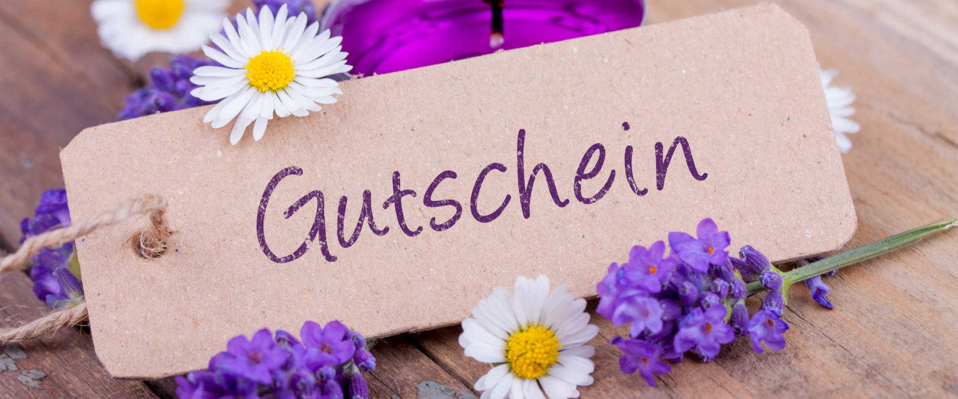Papieranhänger mit der Aufschrift „Gutschein“ zwischen Lavendel und Gänseblümchen auf einem Holztisch